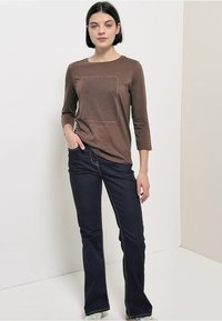 Brun langærmet skjorte med mønstret tekstur, kombineret med mørkeblå vide jeans, stående foran en lys baggrund.