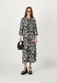 Juoda ir balta raštuota suknelė su ilgomis rankovėmis, juosimi, cinčiuota juosimi ir midi ilgiu. Modelis vilki masyvias juodas platformines sandalus ir juodą rankinę.