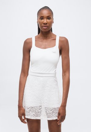 Femeie purtând un top alb fără mâneci și o fustă albă din dantelă, stând în picioare într-un fundal simplu și deschis, privind înainte.