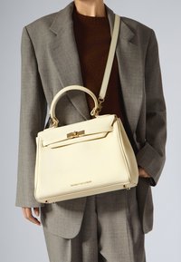 Kremowa skórzana torba na ramię z teksturowaną powierzchnią, górną rączką i regulowanym paskiem crossbody. Wyposażona w złote okucia i logo marki.