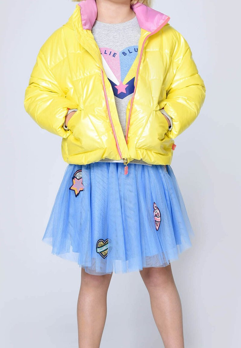 Enfant portant une doudoune jaune brillante avec une doublure rose, un pull graphique gris avec une étoile, et une jupe en tulle bleue avec des patchs colorés.