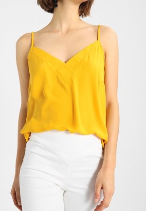 Kvinde iført gul, ærmeløs V-hals top med tynde stropper sammen med hvide, højtaljede bukser mod en ensfarvet baggrund.