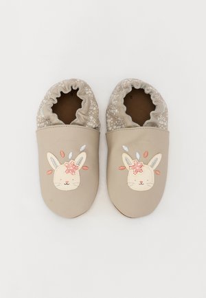 Chaussons pour enfants beiges, avec un bout arrondi, une bordure élastique en motif floral, et un design de visage de lapin mettant en avant une fleur rose et des accents de plumes.