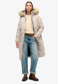 Lys grå lang puffjakke med en faux fur hætte, quiltet tekstur, parret med en cremefarvet striktrøje, blå brede jeans og brune støvler.