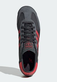 Zapatilla deportiva negra y roja con detalles de ante y texturizados, que cuenta con tres líneas rojas, cordones redondos y una plantilla con patrón.