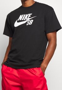 Černé bavlněné tričko s velkým bílým logem "NIKE SB" na přední straně. Krátké rukávy a kulatý výstřih. Hladká textura, volný střih.