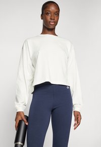 Lys beige cropped sweatshirt med lange ermer og en logo på brystet, sammen med mørkeblå leggings med en logo på hoften.