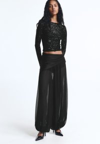 Egy nő fekete flitteres, hosszú ujjú, crop toppot visel, átlátszó, széles lábú fekete nadrágot, amelynek van egy átkelési részlete, valamint fekete, hegyes orrú magassarkú cipőt.