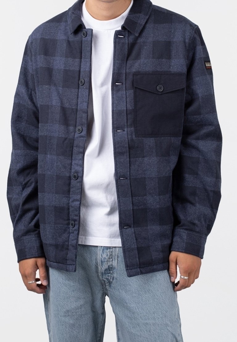 Homme portant une veste à carreaux bleu foncé boutonnée avec une poche poitrine noire unie par-dessus un t-shirt blanc et un jean bleu clair.