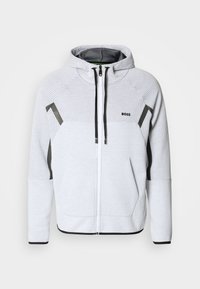 Hoodie zippé gris clair en tissu texturé, avec une capuche, des accents noirs et un logo sur la poitrine à gauche.