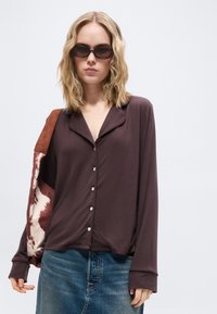 Femme aux cheveux blonds mi-longs portant des lunettes de soleil ovales, une chemise sombre boutonnée, un jean bleu et un sac à épaule marron et blanc à motifs.