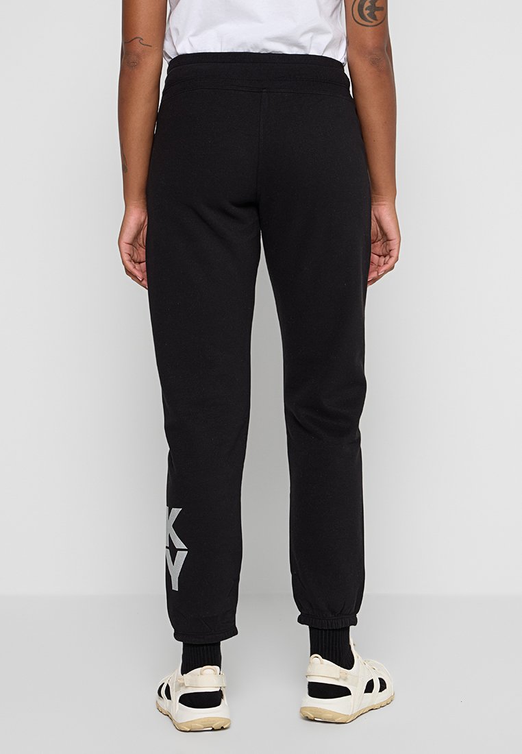 Persona che indossa pantaloni jogger neri con polsini elastici e sneakers bianche, in piedi contro uno sfondo grigio chiaro uniforme, vista da dietro.