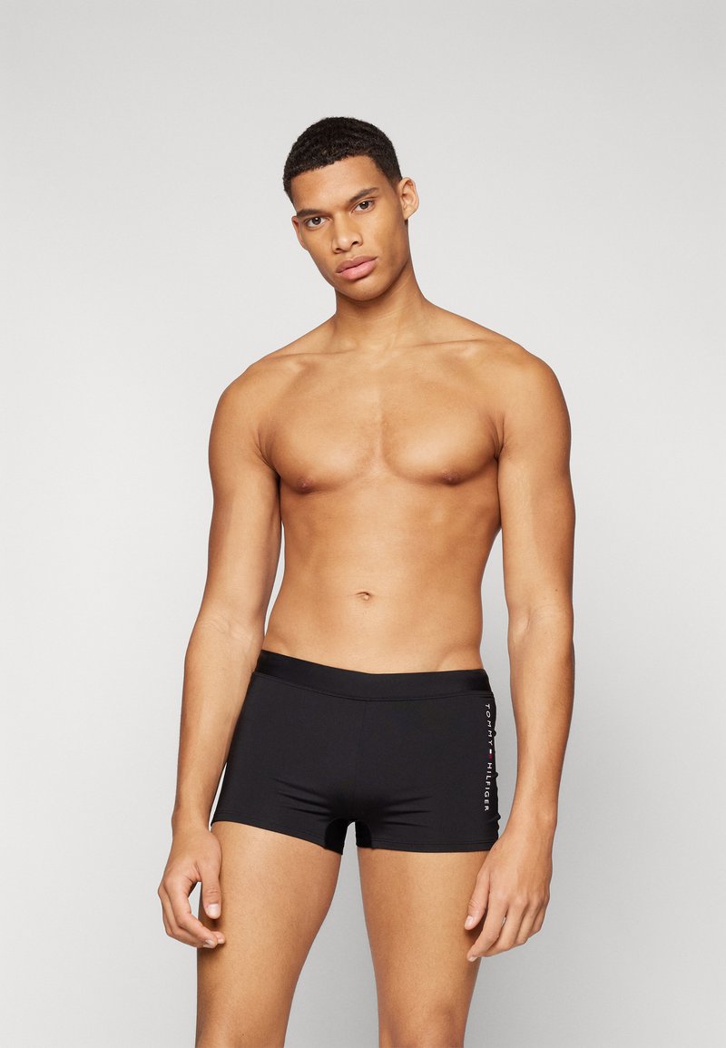 Shorts de bain noirs à coupe ajustée, fabriqués en matériau lisse. Présentent un logo de marque subtil en blanc et rouge sur le côté.