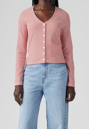 Femme portant un haut à manches longues à rayures horizontales rouges et blanches avec des boutons, et un jean taille haute bleu clair, sur un fond uni.