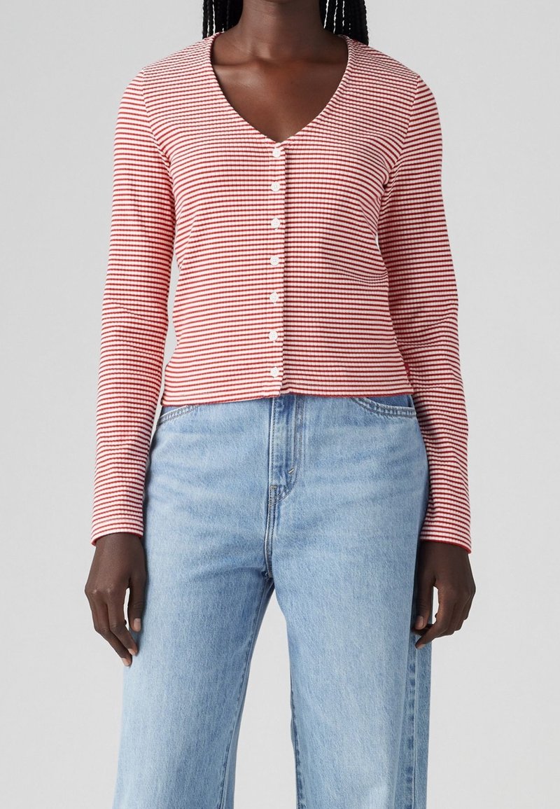 Femme portant un haut à manches longues à rayures horizontales rouges et blanches avec des boutons, et un jean taille haute bleu clair, sur un fond uni.
