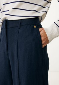 Marineblauwe linnen broek met een soepele textuur en zijzakken; heeft een gouden accent bij de tailleband. Gepaard met een wit gestreept trui.