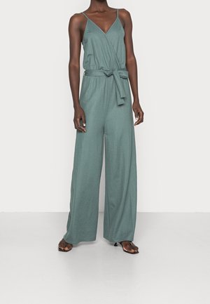 Vrouw draagt een mouwloze groene jumpsuit met V-hals, taillekoord, broek met wijde pijpen en zwarte sandaalhakken, staand tegen een witte achtergrond.