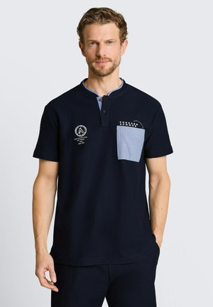 Mann mit hellbraunen Haaren und Bart, der ein marineblaues Kurzarmhemd mit blauer Tasche und dem aufgedruckten Text „Forever Summer“ sowie Logos trägt.