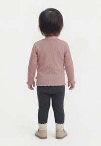 Kisgyermek háttal áll, rózsaszín hosszú ujjú felsőt, sötétszürke leggingset, krémszínű zoknit és bézs cipőt visel fehér háttéren.