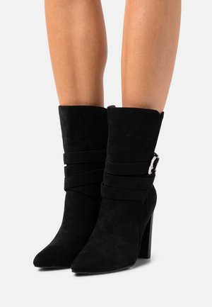 Černé semišové kozačky do půli lýtek se špičatou špičkou, tlustým podpatkem, dekorativními pásky a stříbrnou sponou, nošené na bílém pozadí na holých nohou.