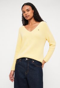 Sárga bordázott kötött pulóver V-nyakkal és hosszú ujjakkal, bal oldalon egy kis logó részlettel. Sötétkék farmernadrággal párosítva.