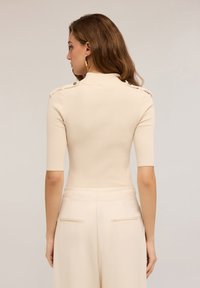 Kobieta z długimi brązowymi włosami, ubrana w beżowy prążkowany top z krótkim rękawem i guzikami na ramionach oraz pasujące spodnie z wysokim stanem, pokazana od tyłu.