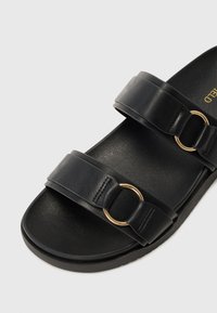 Black sandal featuring two wide straps with a gold circular accent, minimalist design, smooth texture, and flat sole.