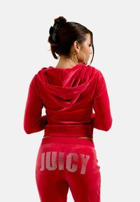 Tuta in velluto rosso con cappuccio; i pantaloni presentano la scritta "JUICY" in strass sul retro. Tessuto liscio, design aderente e polsi a costine.