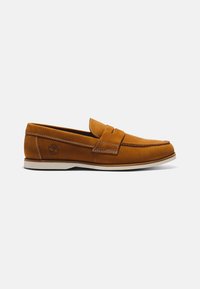 Mocassin en cuir marron à enfiler avec semelle blanche et détails cousus, vue de côté sur un fond clair uni.