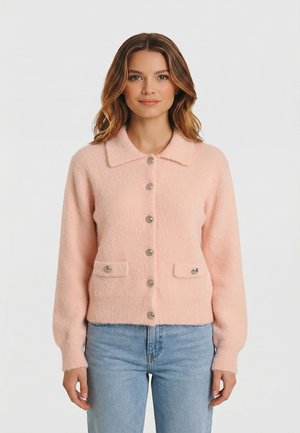 Donna che indossa un cardigan rosa chiaro peloso con bottoni e tasche frontali, abbinato a jeans azzurri, su uno sfondo bianco semplice.