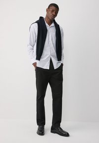 Męski strój składający się z białej koszuli w paski na guziki, granatowego swetra zarzuconego na ramiona, czarnych spodni i czarnych butów.