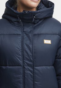 Navy blue puffer jacket with a high collar, zippered front, and quilted segments. Features a small tan logo patch on the chest area.