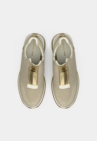 Beiget slip-on tennarit, joissa on teksturoitu verkkopäällinen, kullanvärinen vetoketju, valkoiset yksityiskohdat ja kullanvärinen logo kielen kohdalla.