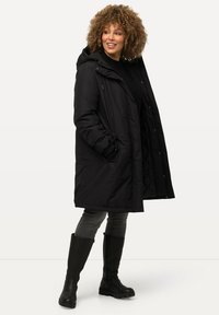 Doudoune noire avec capuche, dotée d'une fermeture éclair et de boutons, de poches latérales et d'un intérieur matelassé. Portée avec un jean gris et des bottes noires montantes.