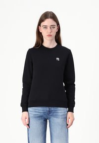 Felpa nera in cotone con maniche lunghe, collo rotondo e polsini a coste. Presenta un piccolo logo bianco sul petto. Colore uniforme con texture liscia.