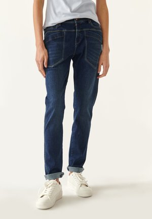 Džíny z denimu v tmavě modré barvě, slim fit s kontrastním prošíváním. Obsahují pět kapes, ohrnuté manžety a jemné blednutí podél předních nohavic.