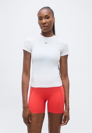 Femeie în picioare, purtând un top alb Nike cu mâneci scurte și pantaloni scurți atletici roșii vii, potriviți, pe un fundal simplu, deschis.