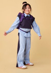 Ragazzina sorridente, vestita con una maglietta a righe blu e pantaloni con un giubbotto blu scuro e uno zaino viola a fantasia rosa.