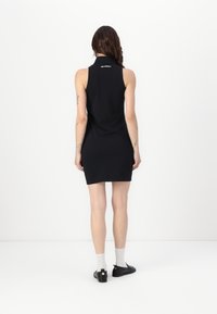 Vestido negro sin mangas con cuello alto, hecho de una tela suave, que cuenta con un detalle de logo en la parte trasera. La prenda se complementa con zapatos negros tipo slip-on y calcetines claros.