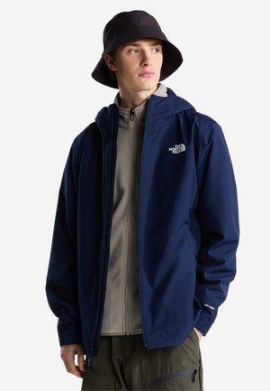 Nuori mies, joka pukeutunut laivastonsiniseen The North Face -takkiin, harmaaseen vetoketjufleeceen, vihreisiin housuihin ja mustaan kalastajahattuun, katsoo oikealle.
