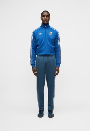 Férfi áll szemben előre nézve, kék Adidas melegítőfelsőt és nadrágot visel, fehér csíkokkal az ujjakon és a lábakon, egyszínű háttér előtt.