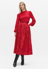 Czerwony top z długim rękawem w połączeniu z czerwoną spódnicą o długości do połowy łydki, ozdobioną koronką w kwiaty. Modelka ma na sobie czarne kozaki za kolano.