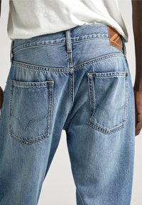 Ljusblå denimjeans med avslappnad passform, två bakfickor med sömdekor, och en brun läderlogotyp på midjebandet.