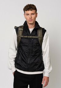 Gilet nero con zip, dal texture lucido e trapuntato. Presenta un colletto alto e due tasche laterali, indossato sopra una camicia a maniche lunghe di colore chiaro.