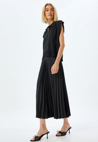 Falda plisada negra con un diseño de cintura alta, combinada con una blusa negra sin mangas y sin cuello. Modelo lleva tacones negros de abertura en los dedos.