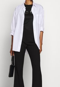 Camisa blanca de manga larga con botones sobre una camiseta gráfica negra, combinada con pantalones acampanados negros y un pequeño bolso negro.