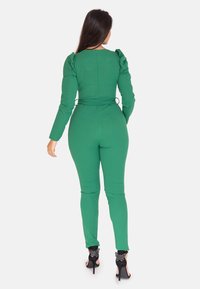 Grön jumpsuit med långa ärmar, figurnära silhuett, texturerat tyg och dragkedja bak. Höga klackskor med fjärilsdetaljer.