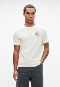 Cremefarvet bomulds-T-shirt med rund udskæring og korte ærmer, prydet med et cirkulært logo i rød og marineblå på venstre bryst.