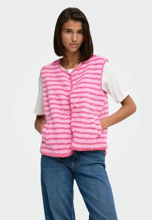 Femme portant un gilet duveteux rayé rose et rose clair par-dessus un t-shirt blanc à manches courtes et un jean bleu, les mains dans les poches du gilet.