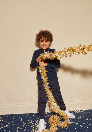 Bambino sorridente con capelli ricci tiene una ghirlanda di stelle d’oro e d’argento, in piedi su un tappeto blu con coriandoli d’oro e bianchi sparsi.